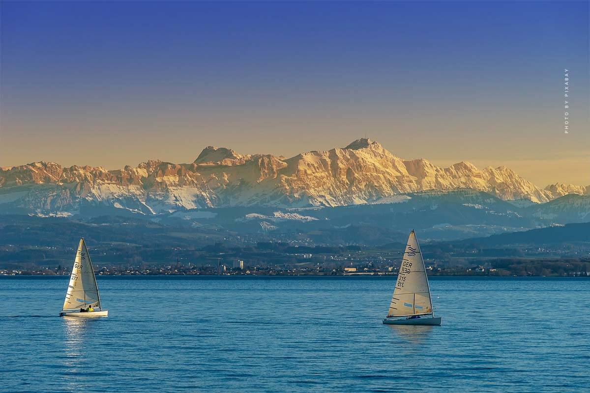Des bateaux blancs naviguent sur l’eau bleue du lac et les montagnes sont visibles en arrière-plan.