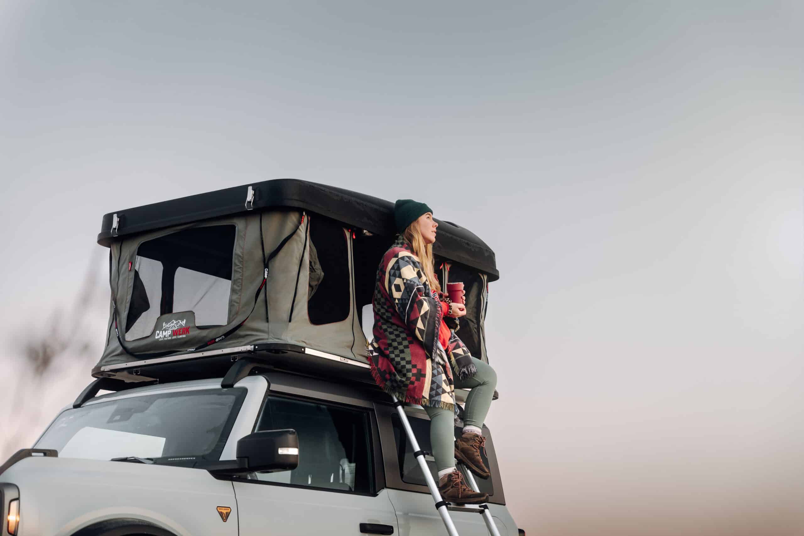 Une femme est assise à l'entrée de sa tente de toit et profite de la nature. Il y a assez de place pour cela dans la Good Vibes, car elle offre une hauteur sous plafond étonnamment importante. La Good Vibes est la première tente de toit à coque rigide à ouverture verticale de Campwerk.