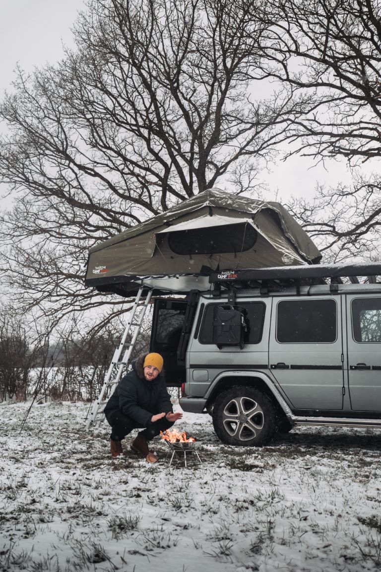 Un campeur se réchauffe les mains au feu à côté de sa voiture avec une tente de toit dans la neige