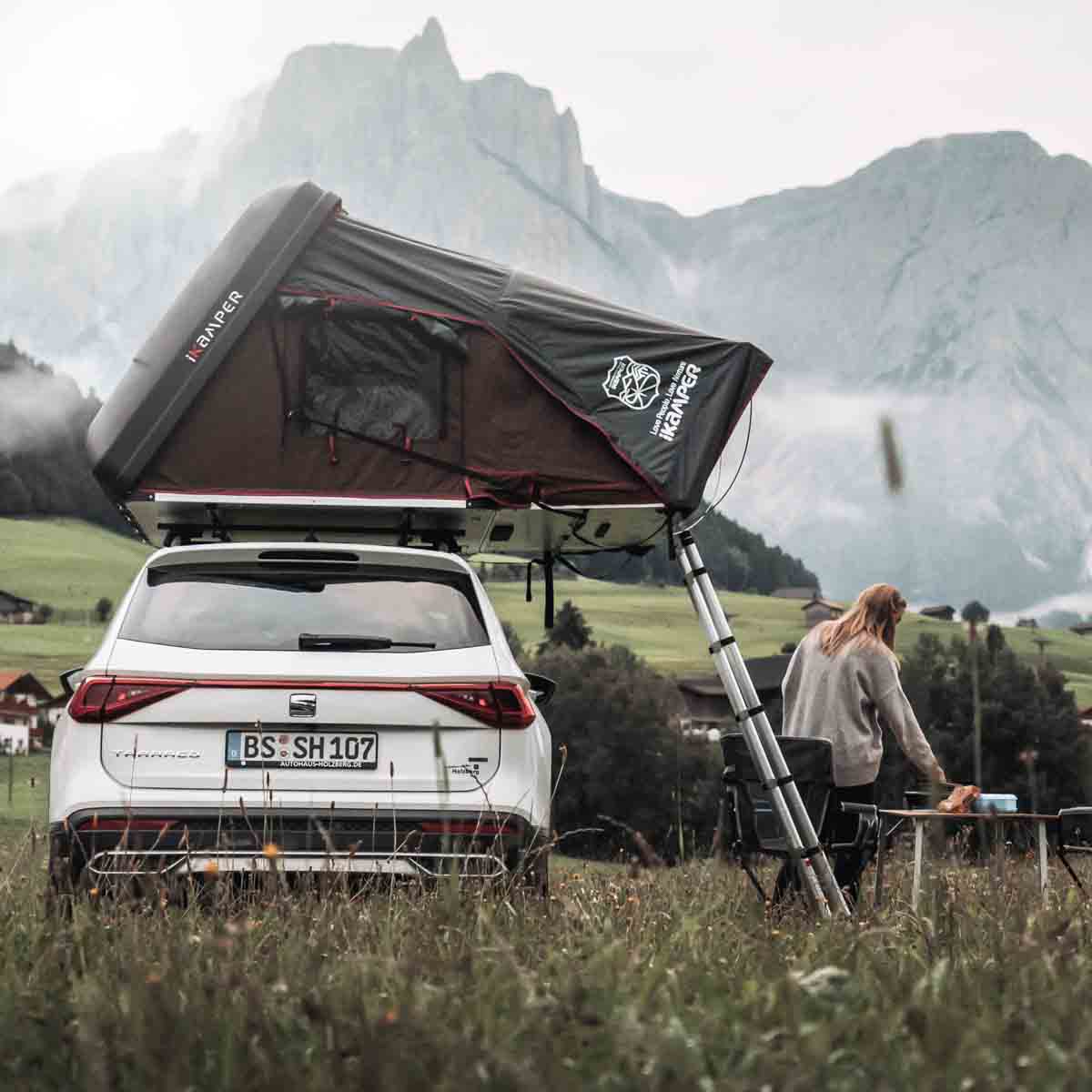 Une femme se tient dans les montagnes à côté de sa voiture avec une tente de toit