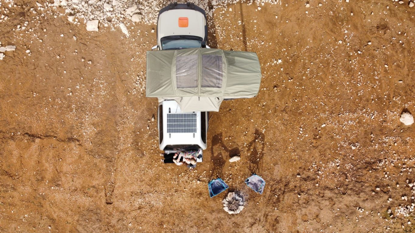 Tente de toit Adventure de Campwerk vue de dessus. Un couple est assis à l'arrière de la voiture, à côté se trouvent des chaises de camping et un foyer 