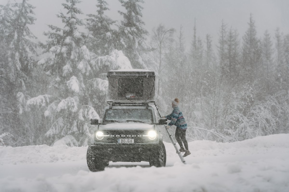 Une femme monte à l'échelle de sa tente de toit à coque rigide CAMPWERK lors d'une tempête de neige