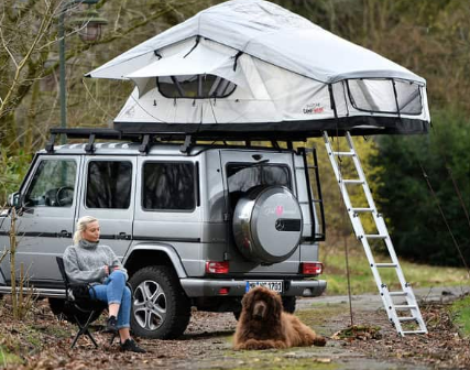 Une femme assise sur une chaise de camping avec son chien devant sa voiture avec une tente de toit Adventure