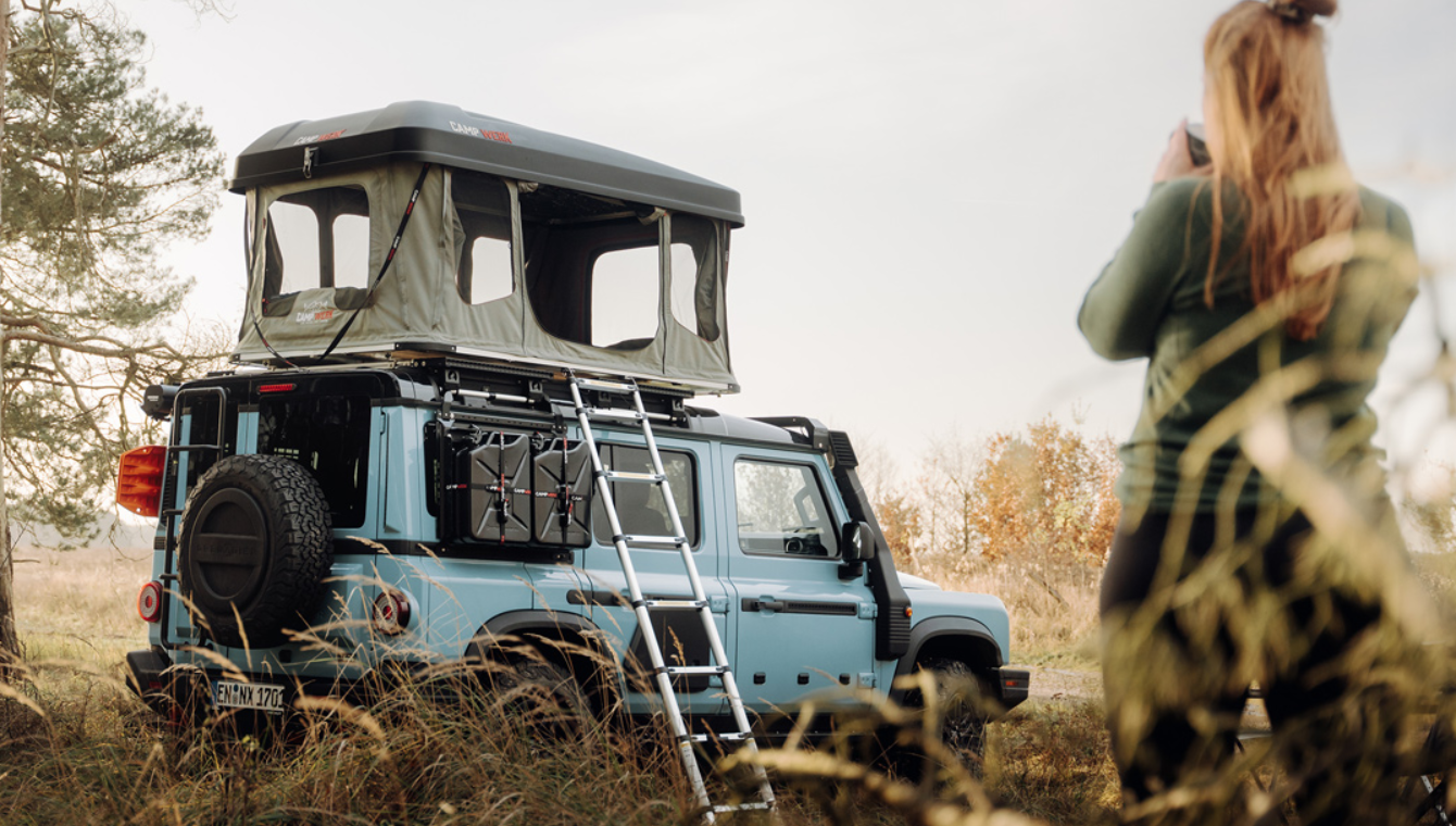 Une femme boit une tasse de café devant sa voiture avec une tente de toit Good Vibes